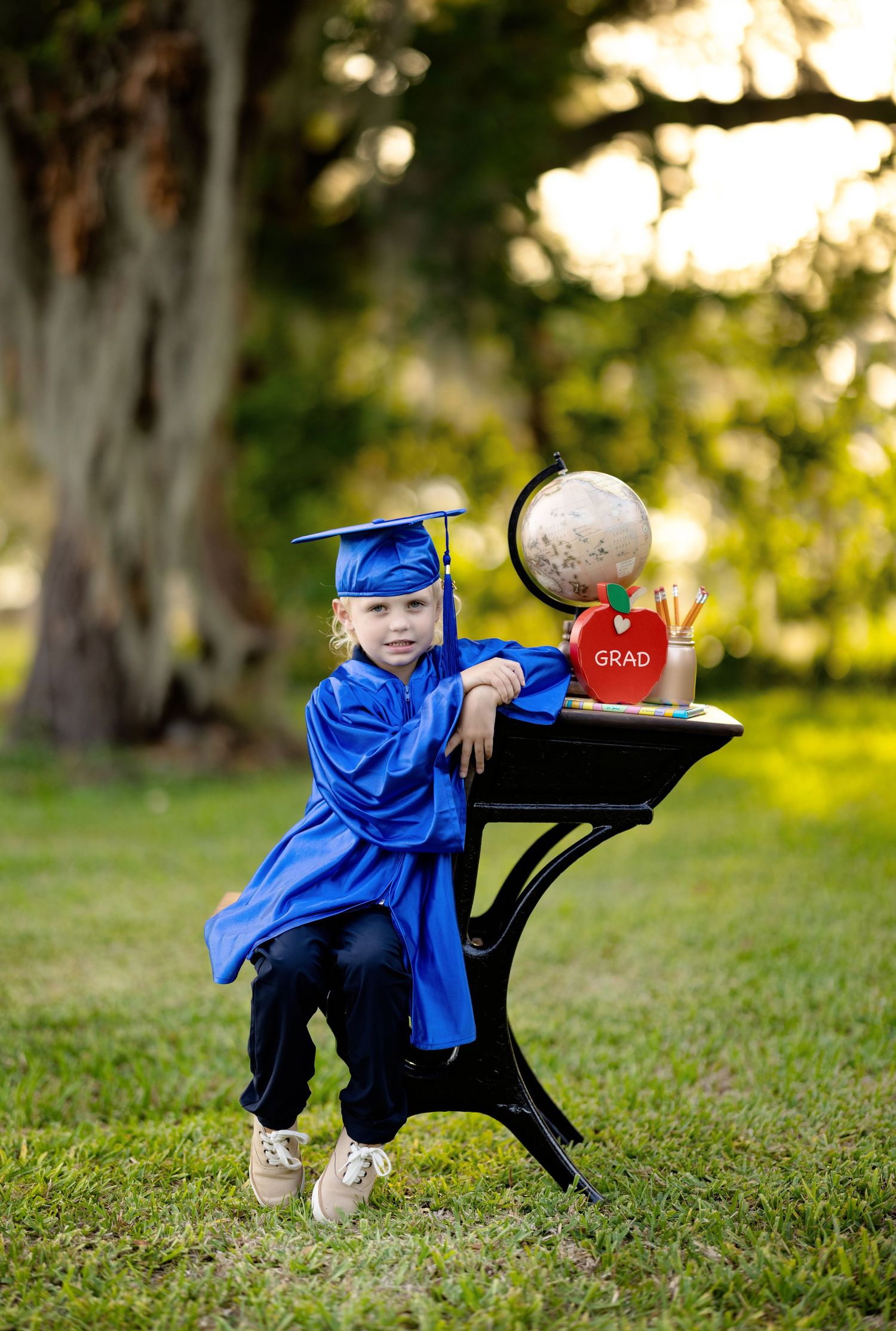 Young child in blue graduation cap and gown poses at school desk outdoors with globe and GRAD apple.