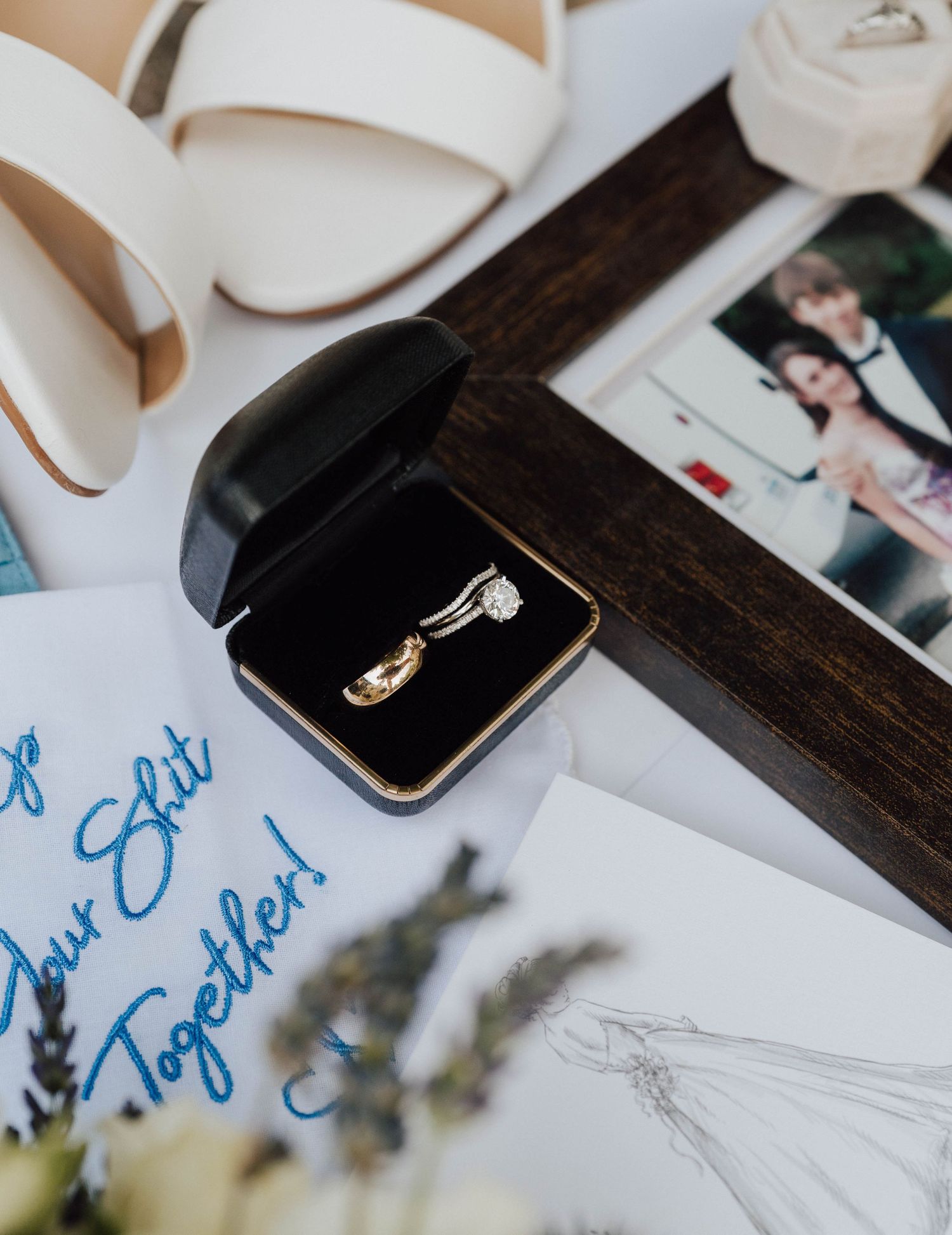 Gold wedding rings in black box beside bridal shoes, photo, dress sketch, and blue handkerchief on white surface.