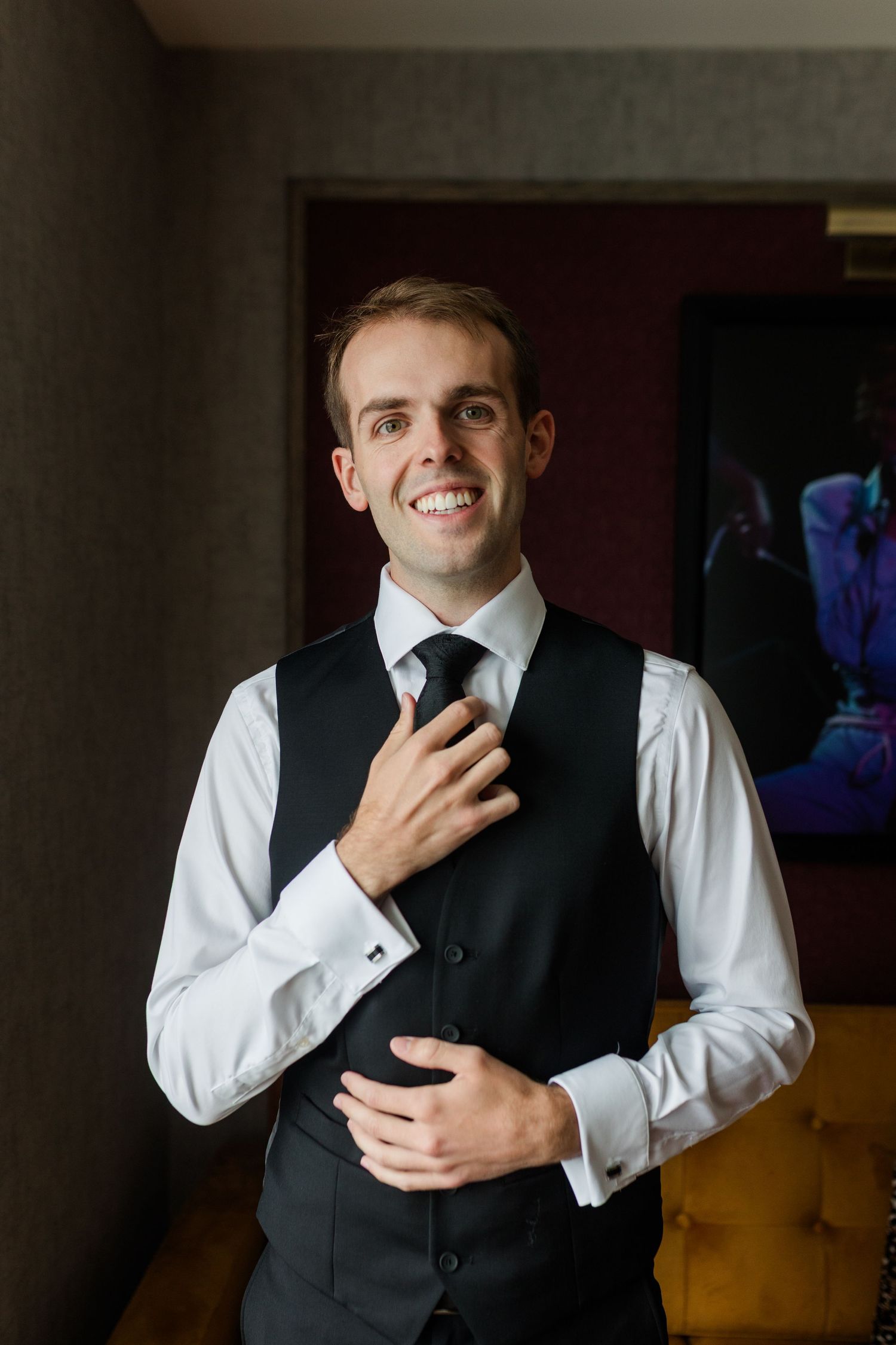 Smiling groom in white dress shirt, black vest and tie adjusting his necktie while getting ready for wedding.