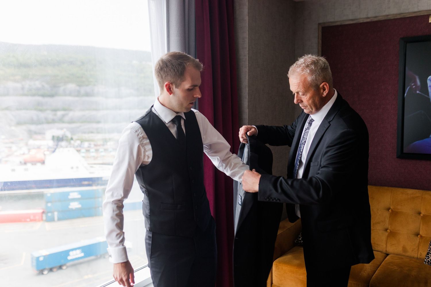 Two men in formal attire helping each other prepare for a wedding near a large window with harbor view.