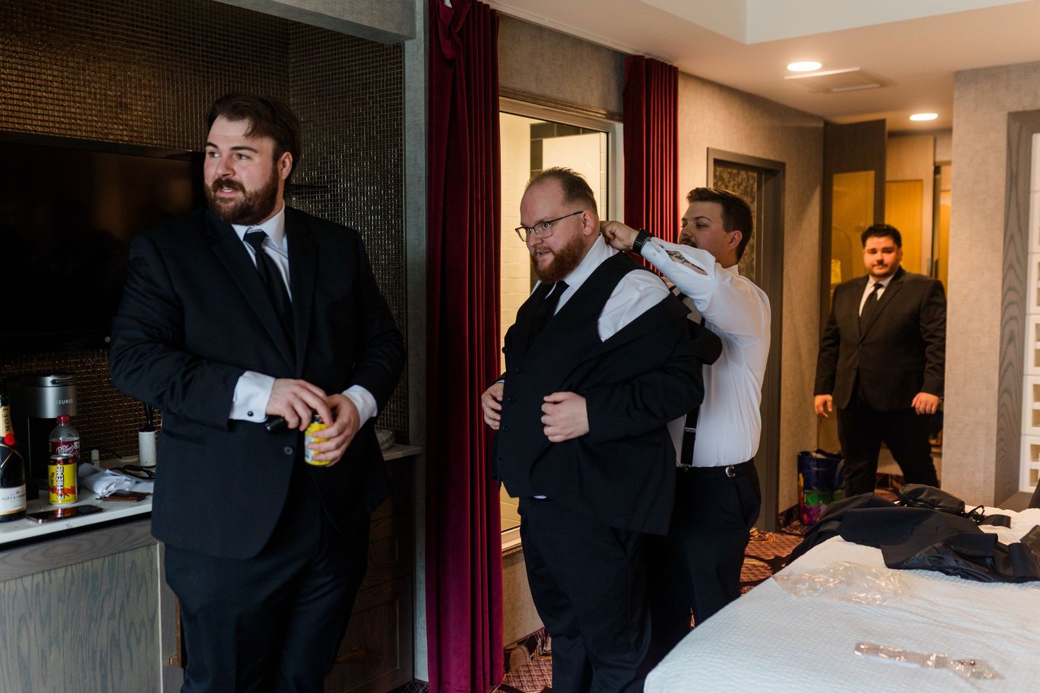 Groomsmen in black suits help each other get dressed in a hotel room before a wedding ceremony.