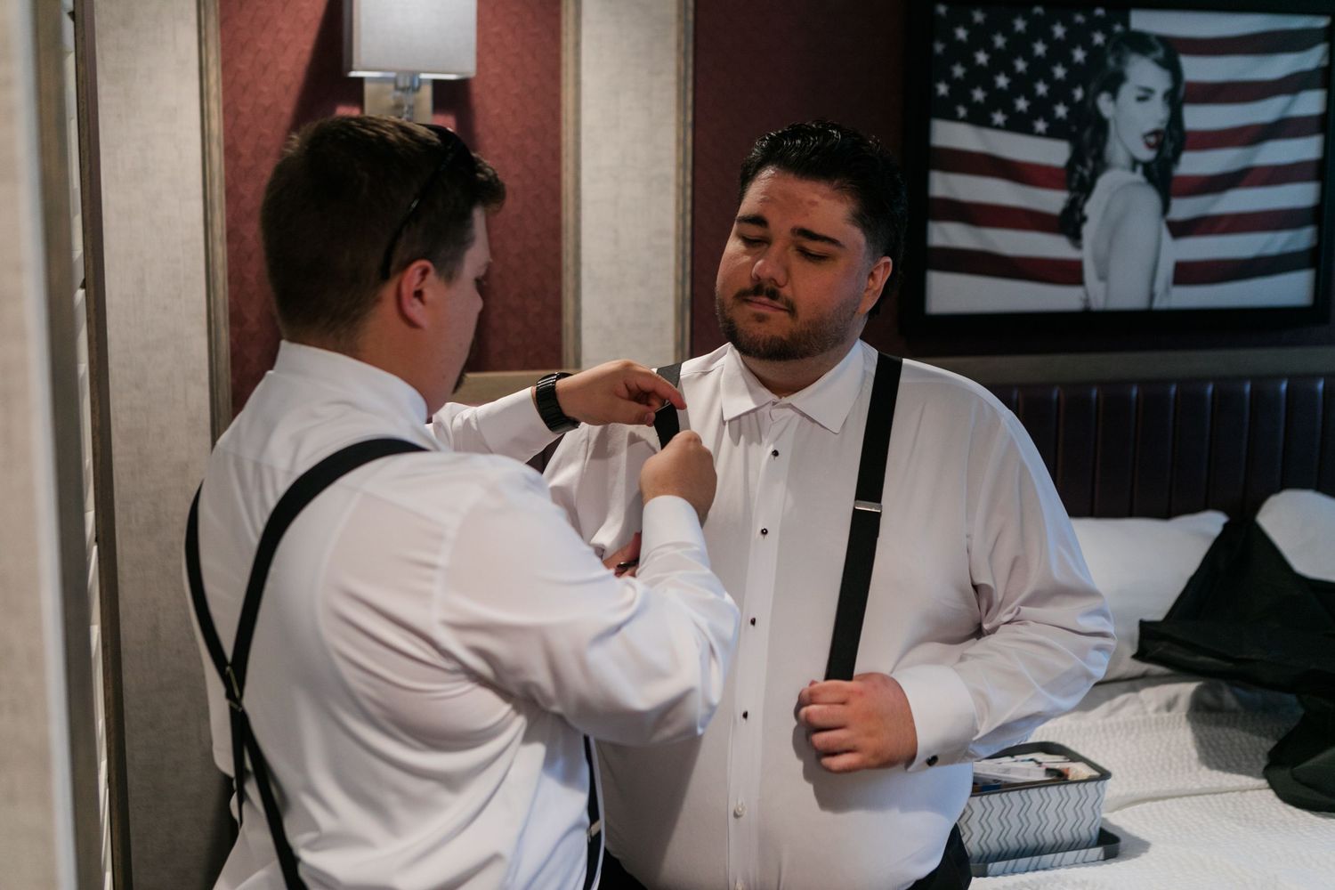 Two men in white dress shirts and suspenders help each other get ready for a formal event in a hotel room.