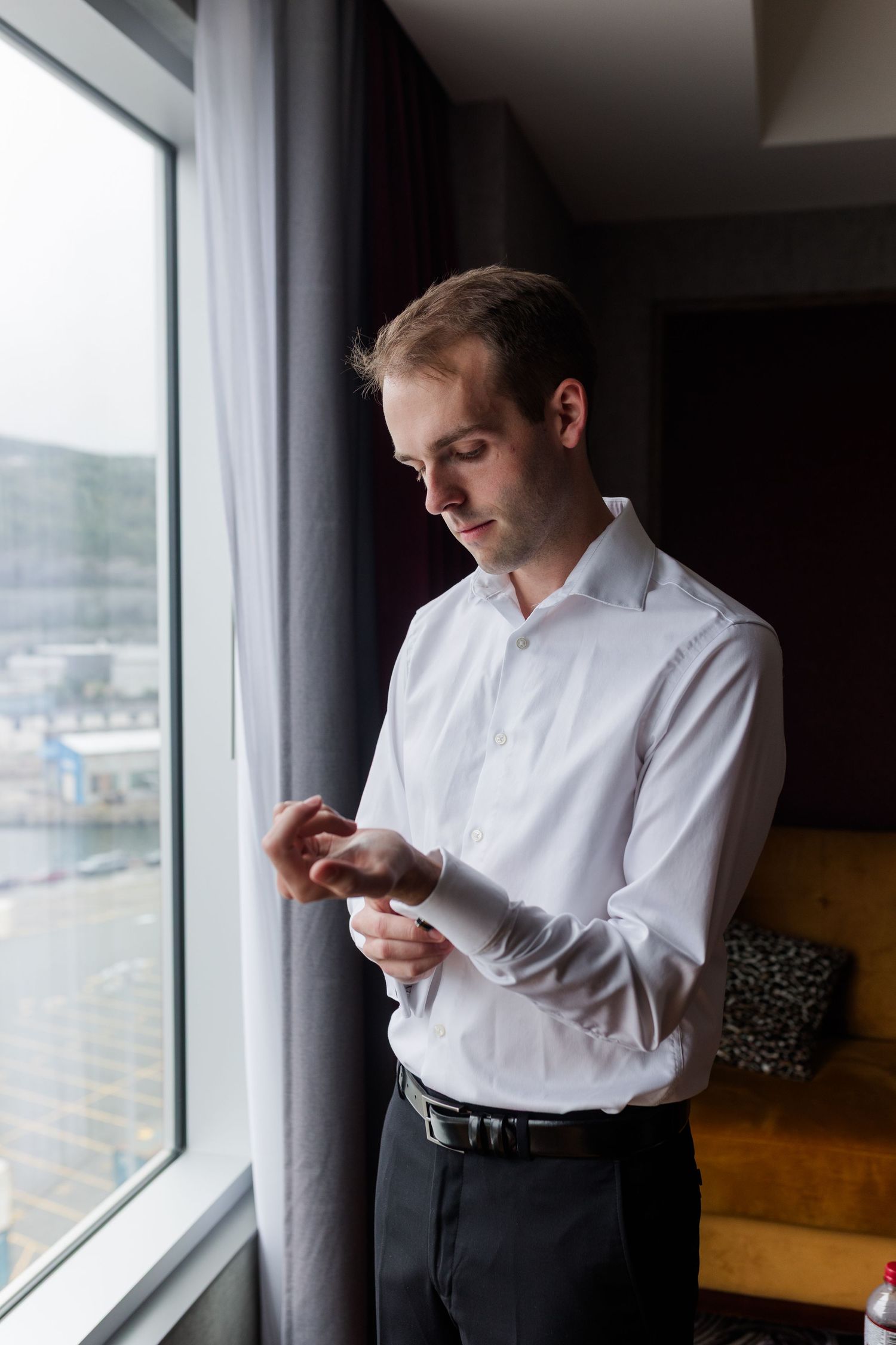 Man in white dress shirt buttoning his cuff link while standing by a window in a hotel room.