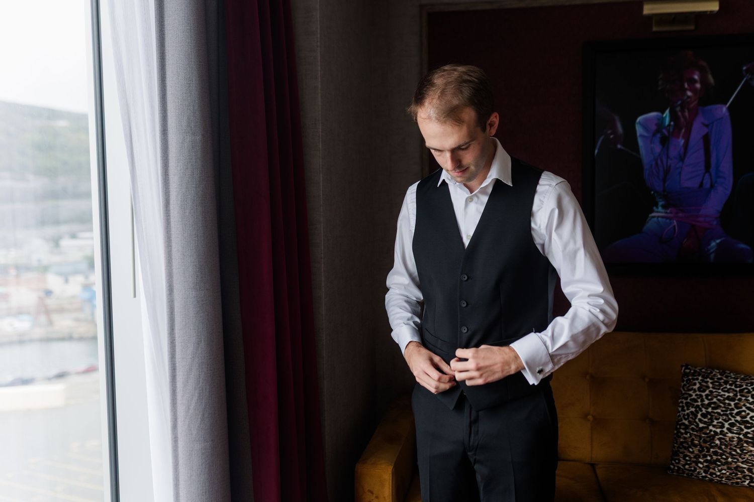 Groom in black vest and white shirt buttoning up near a window before wedding ceremony.