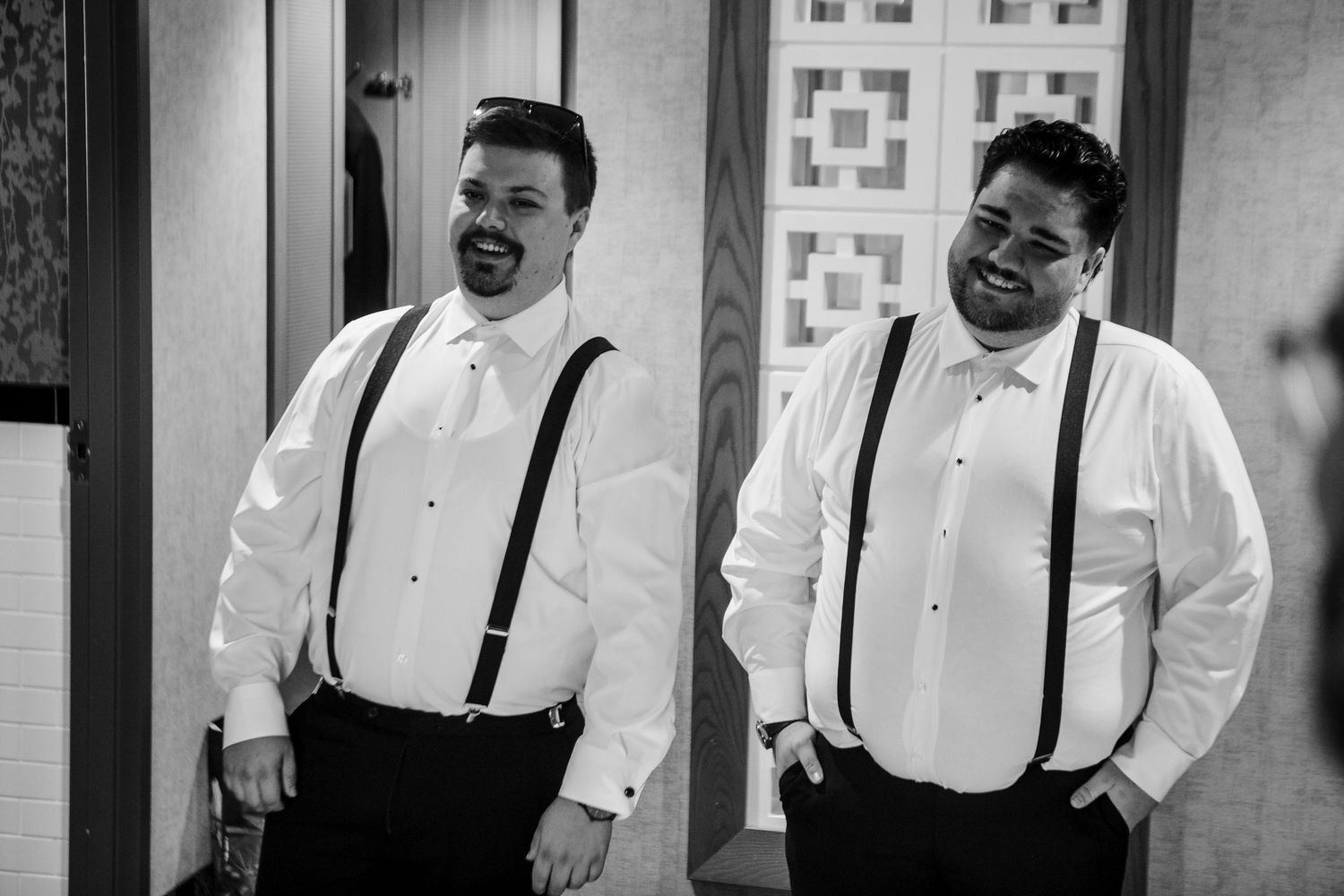 Two groomsmen in white shirts and suspenders laughing together before a wedding ceremony in black and white photo.