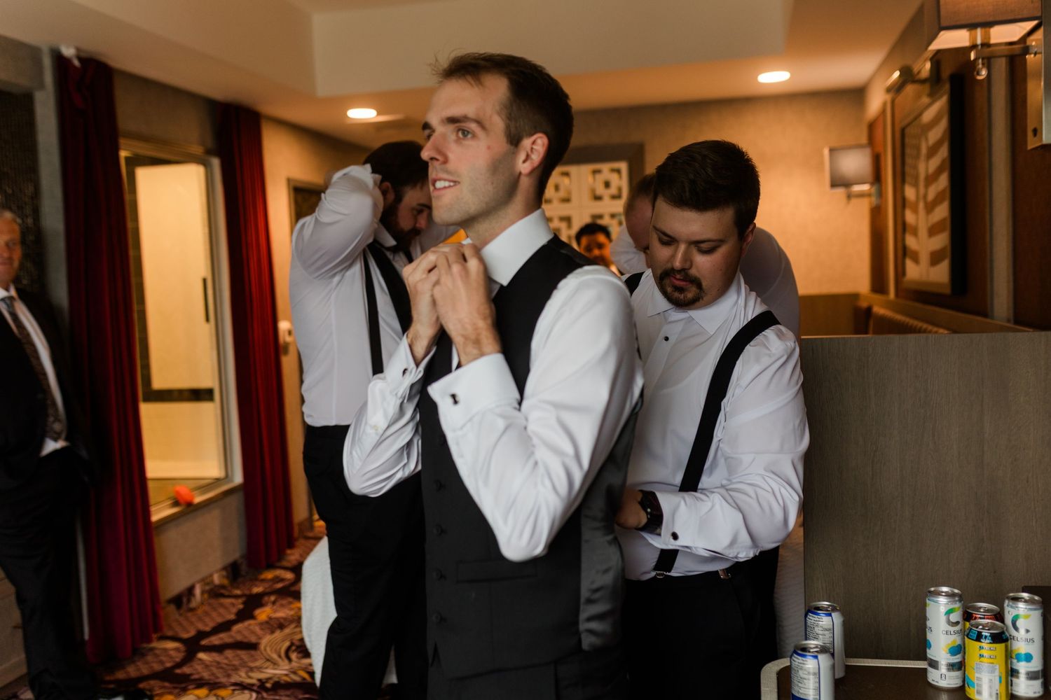 Groomsmen helping groom adjust tie and suspenders while getting ready for wedding ceremony indoors.