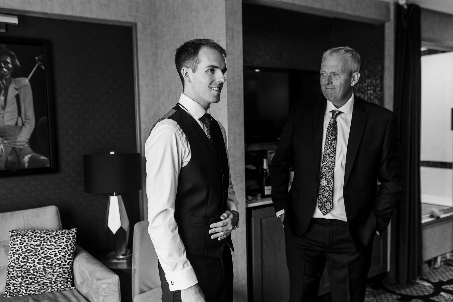 Black and white photo of two men in formal attire conversing in a hotel room, likely a groom and father before a wedding.