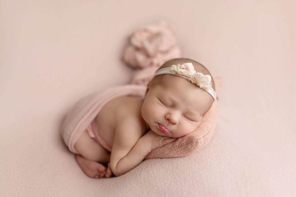 Sleeping newborn baby girl in pink wrap and floral headband on soft pink backdrop.