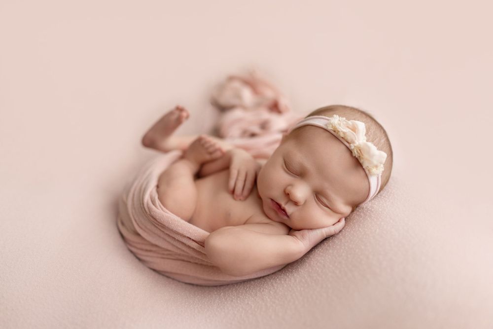 Sleeping newborn baby girl wrapped in pink swaddle with floral headband on soft pink backdrop.