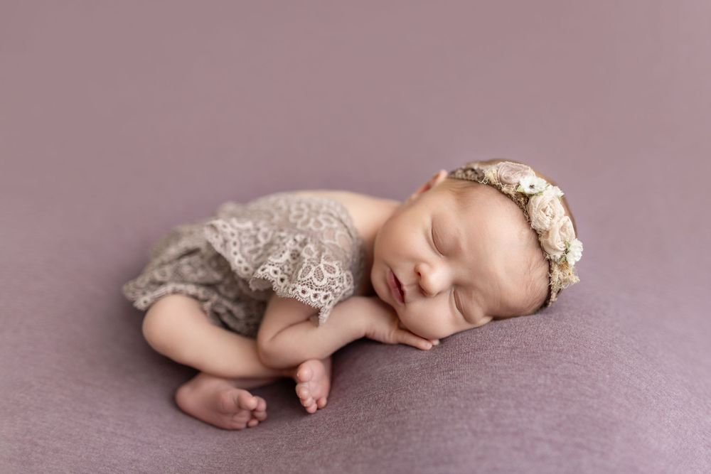 Sleeping newborn baby girl in lace romper and floral headband on purple backdrop.