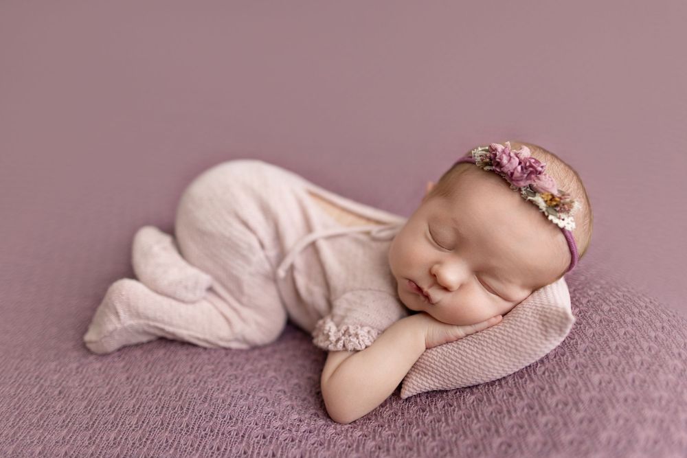 Newborn baby girl sleeping peacefully in pink romper and floral headband on lavender backdrop.