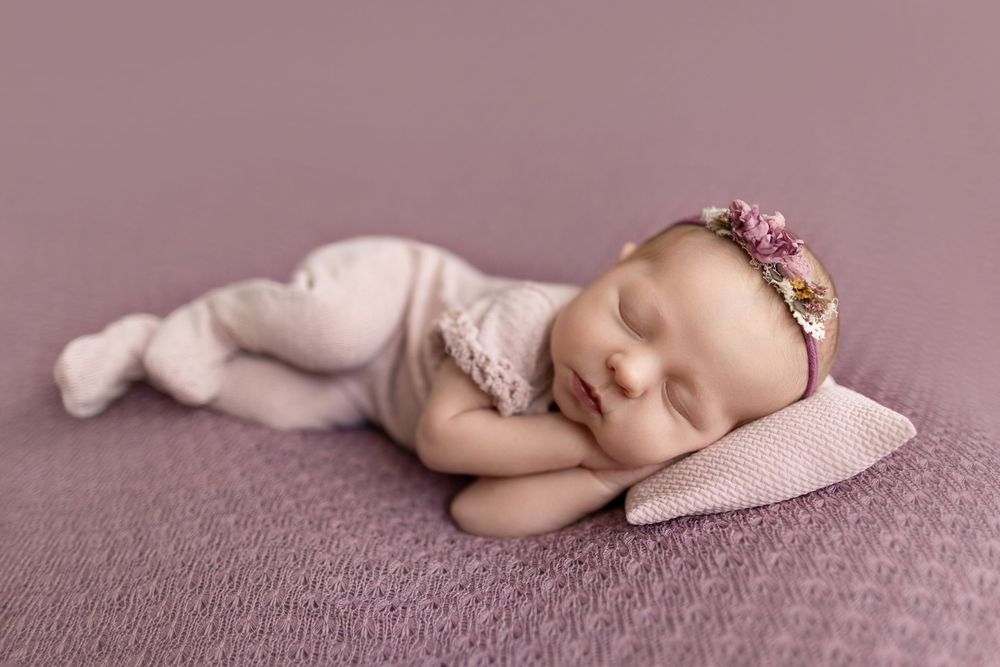 Newborn baby girl sleeping peacefully on a lavender pillow, wearing a pink outfit and floral headband.