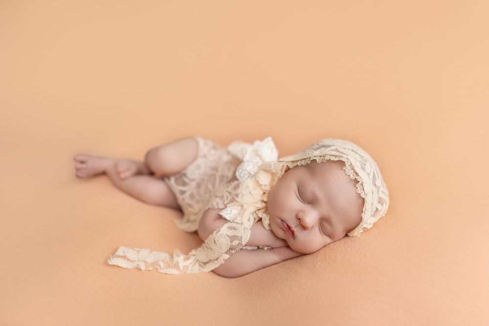 Sleeping newborn baby in cream lace romper and bonnet on peach background.