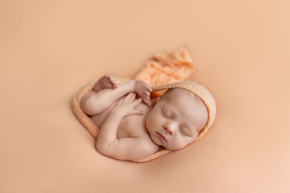 Sleeping newborn baby in peach bonnet curled on matching orange blanket in professional studio photo.