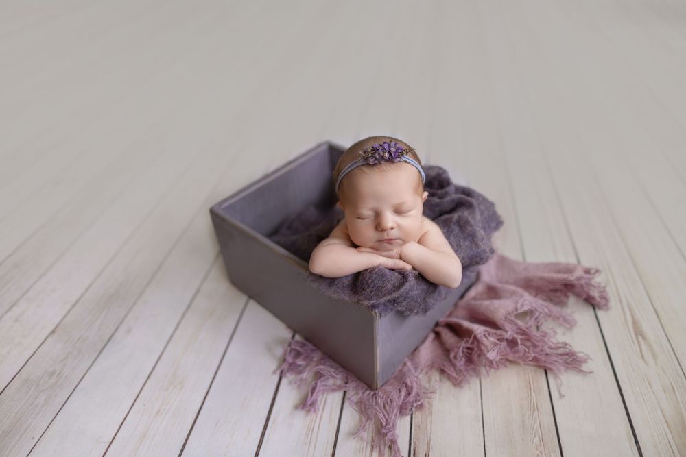 Newborn baby girl sleeping peacefully in a gray wooden box with purple wrap and floral headband.