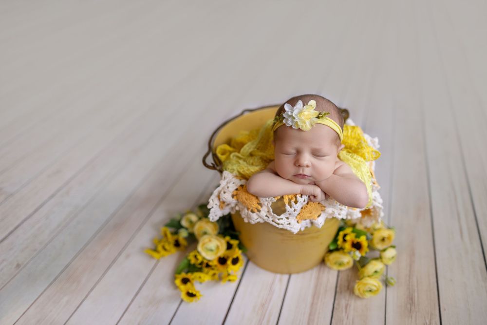 Sleeping newborn baby in yellow bucket with lace, wearing floral headband, surrounded by yellow flowers on white wood floor.