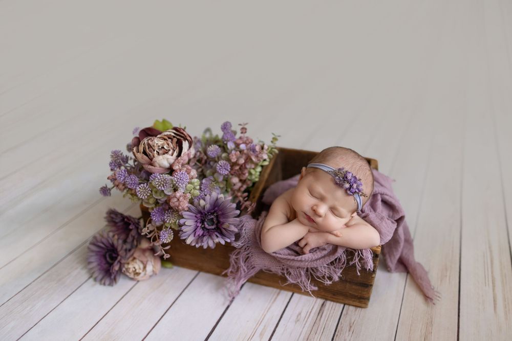 Newborn baby sleeping in wooden box with purple floral arrangement and lavender wrap and headband.