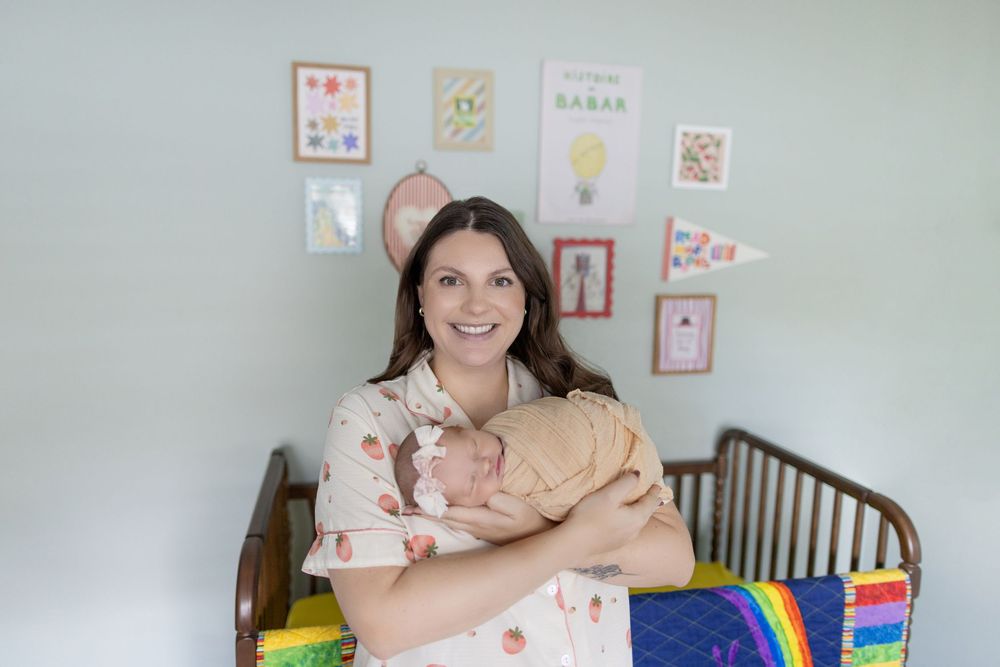 A smiling woman holds a newborn baby in a pastel nursery with a wooden crib and colorful wall art.