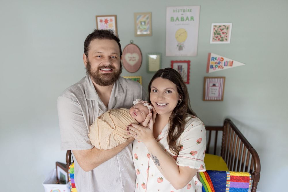 Smiling couple holding their newborn baby in a decorated nursery with colorful artwork on the wall.