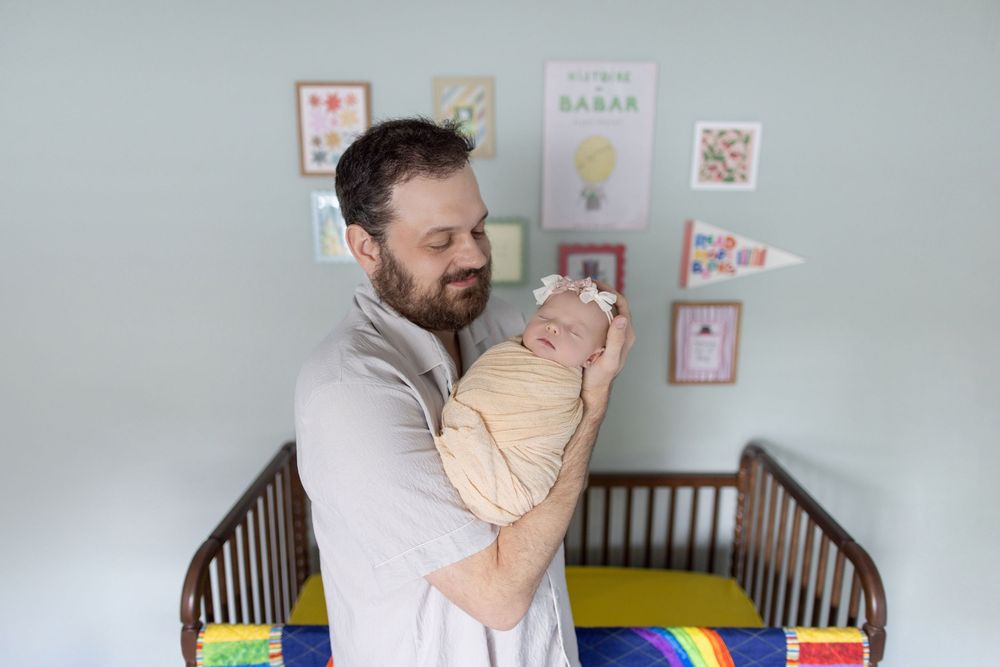 A bearded father tenderly holds his swaddled newborn baby in a decorated nursery with a wooden crib.