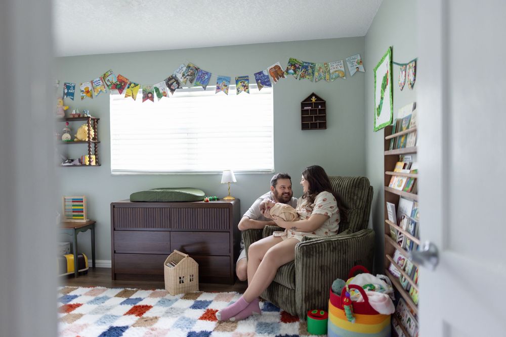 Parents sitting in a rocking chair holding their newborn baby in a colorfully decorated nursery room.