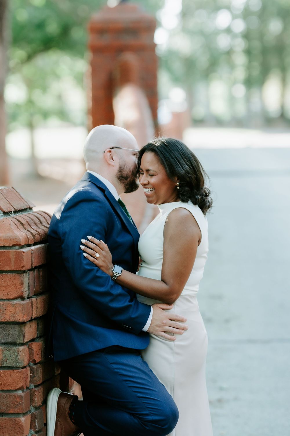 A couple shares a romantic moment leaning against a brick wall outdoors, the woman in white dress laughing joyfully.