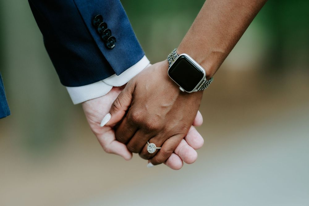 Close-up of an interracial couple holding hands, showcasing a diamond engagement ring and a smartwatch.