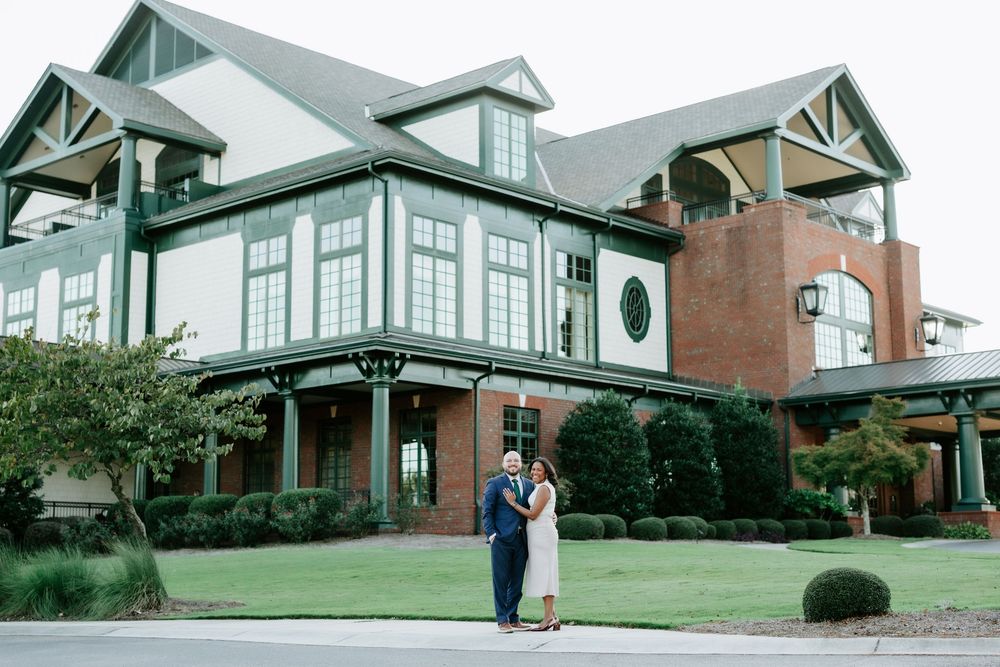 Couple embraces in front of elegant green and brick venue building surrounded by manicured landscaping.