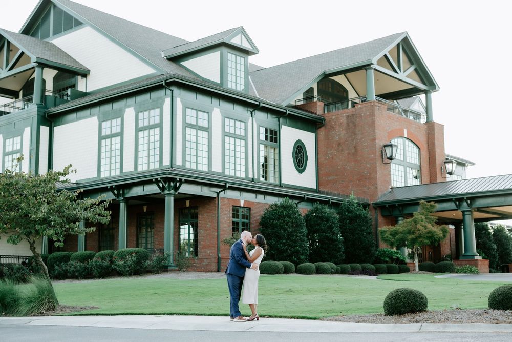 Couple shares a romantic kiss in front of an elegant green and white venue surrounded by lush landscaping.