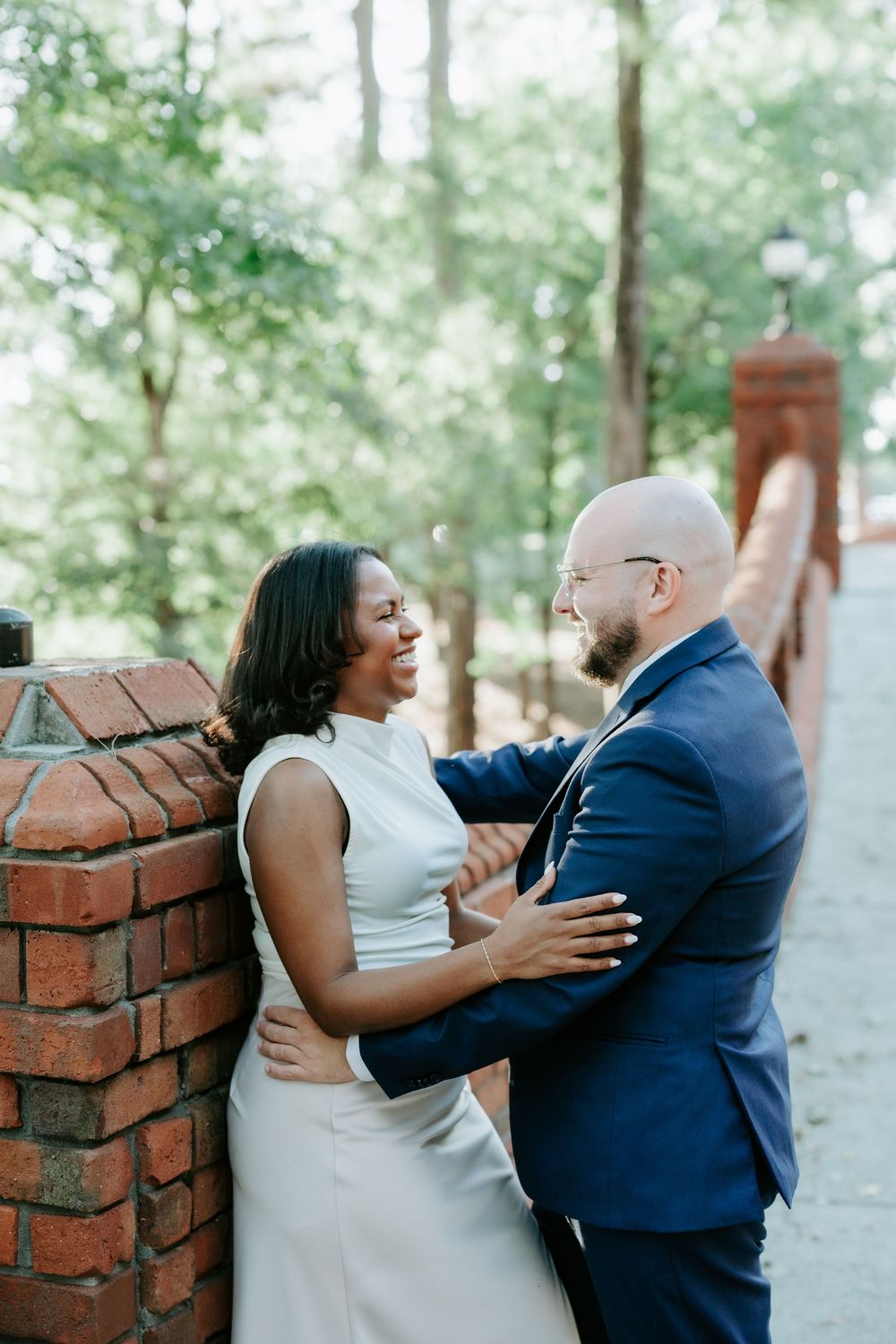 A couple shares a joyful moment on a brick bridge, the woman in white and man in a navy suit embracing outdoors.