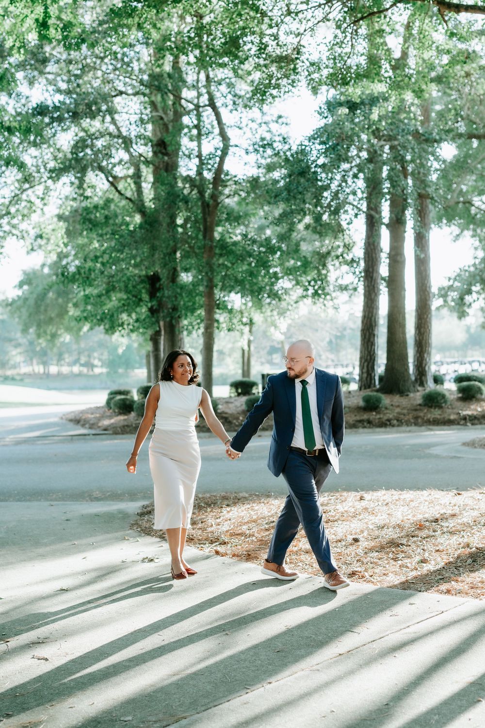 Couple holding hands walking outdoors in formal attire, white dress and navy suit, surrounded by lush green trees.