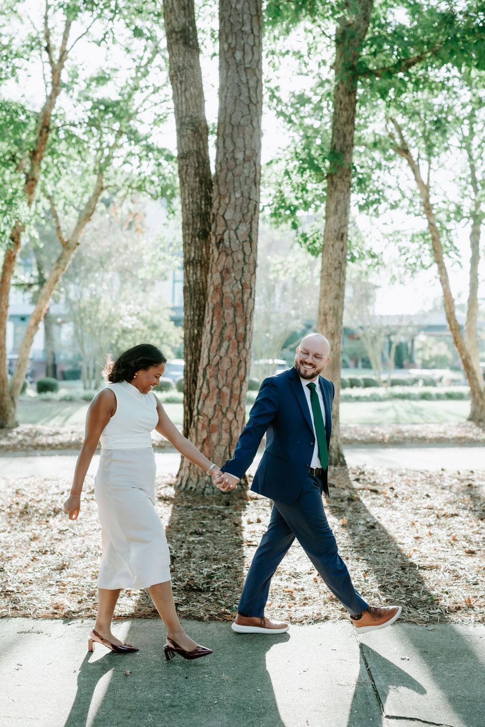A couple holds hands walking through a sunlit park, the woman in a white dress and the man in a navy suit with green tie.