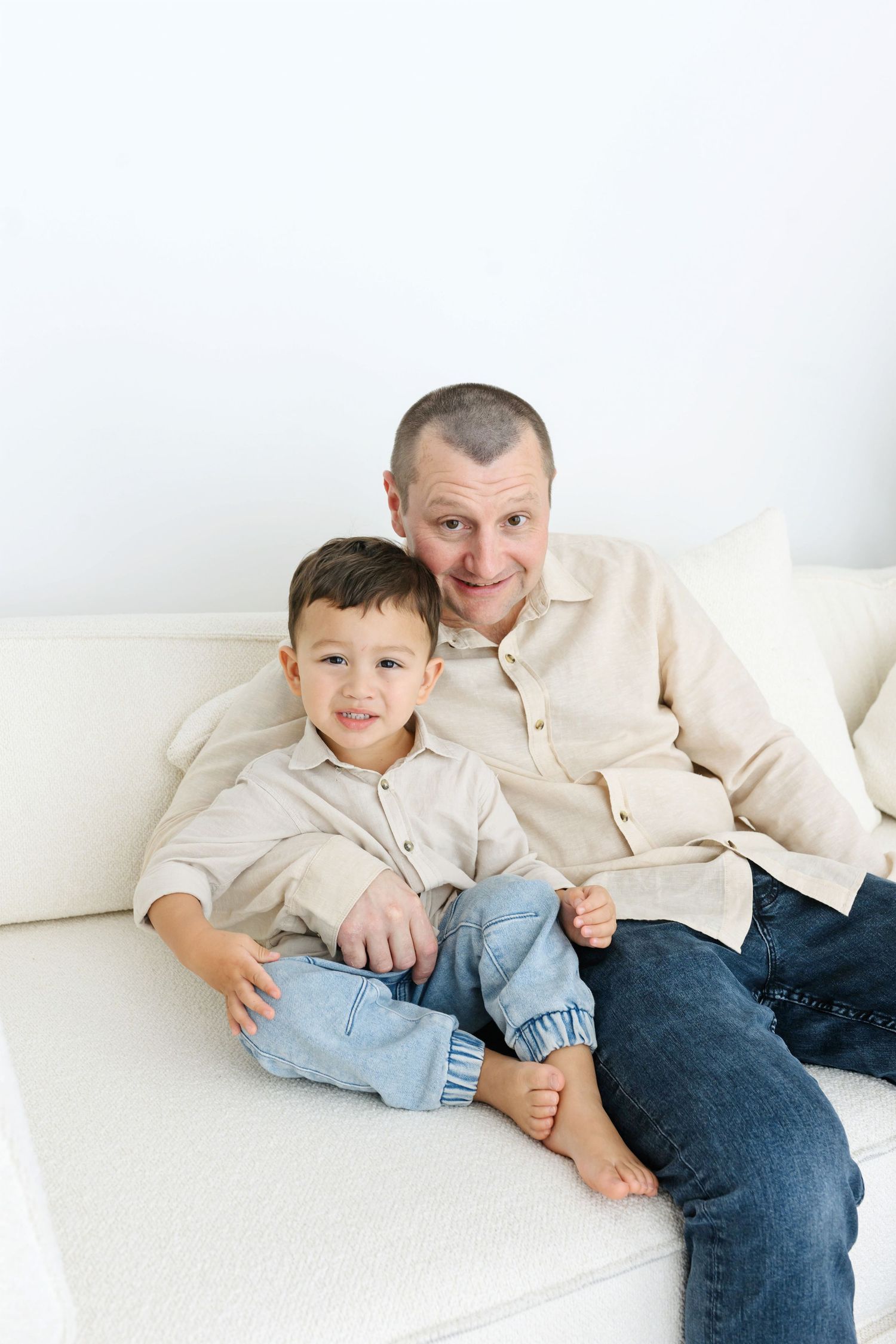 A smiling father and young son sitting together on a white sofa, both wearing beige shirts and jeans.