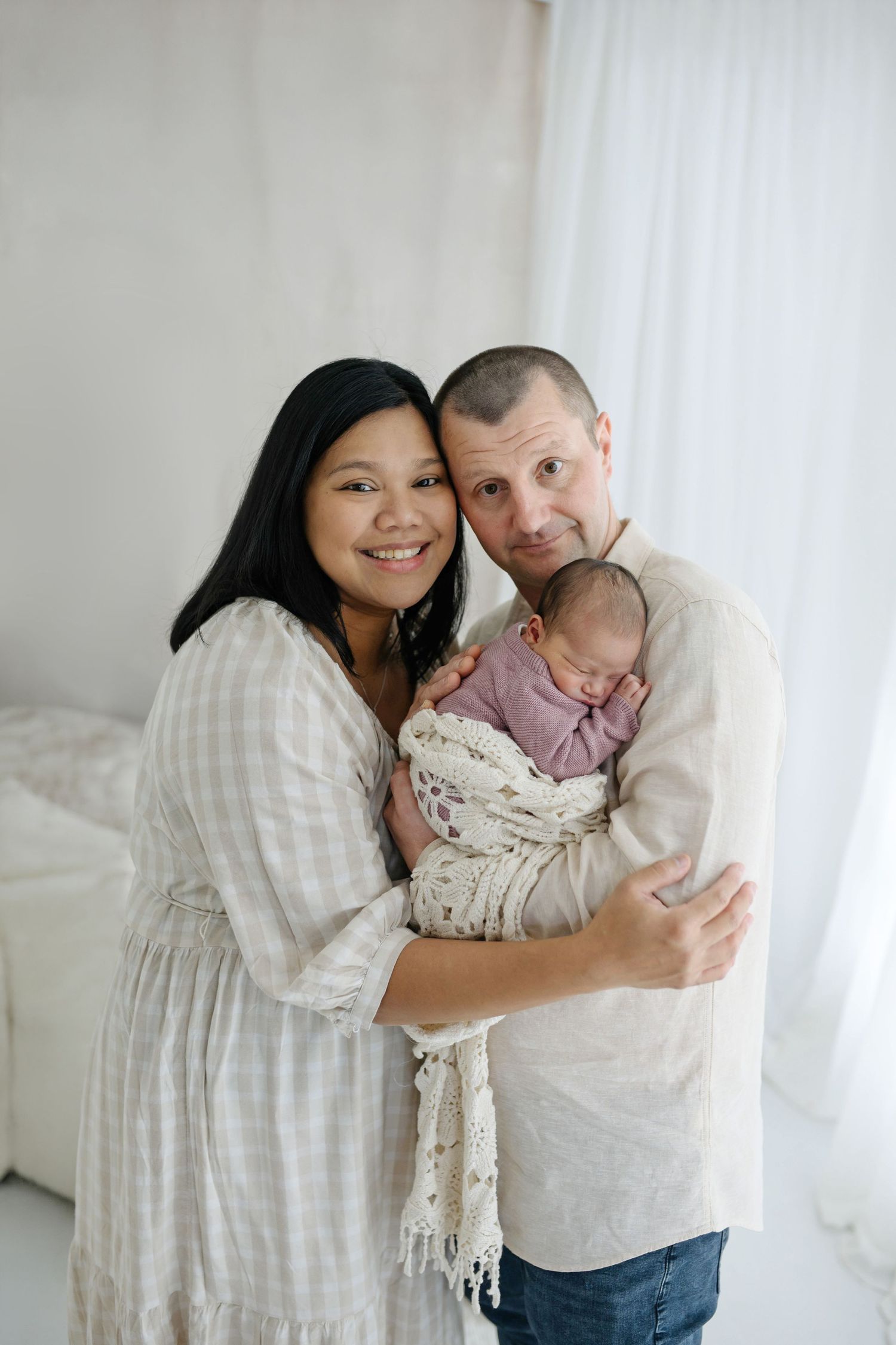 Happy couple in neutral linen clothing holding their swaddled newborn baby in a bright white studio setting.