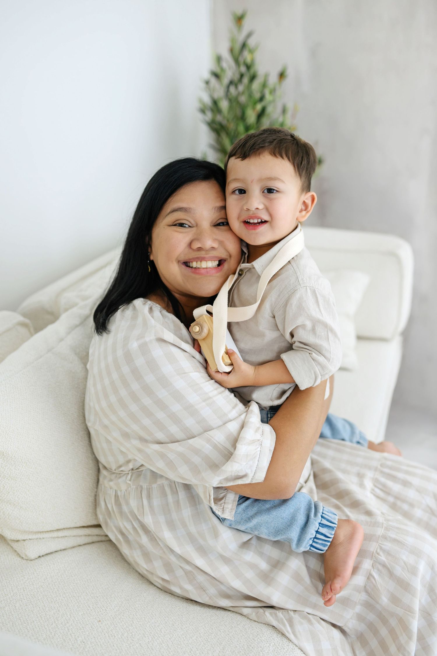 A smiling mother hugs her happy toddler son on a white sofa, both wearing neutral tones in a bright room.