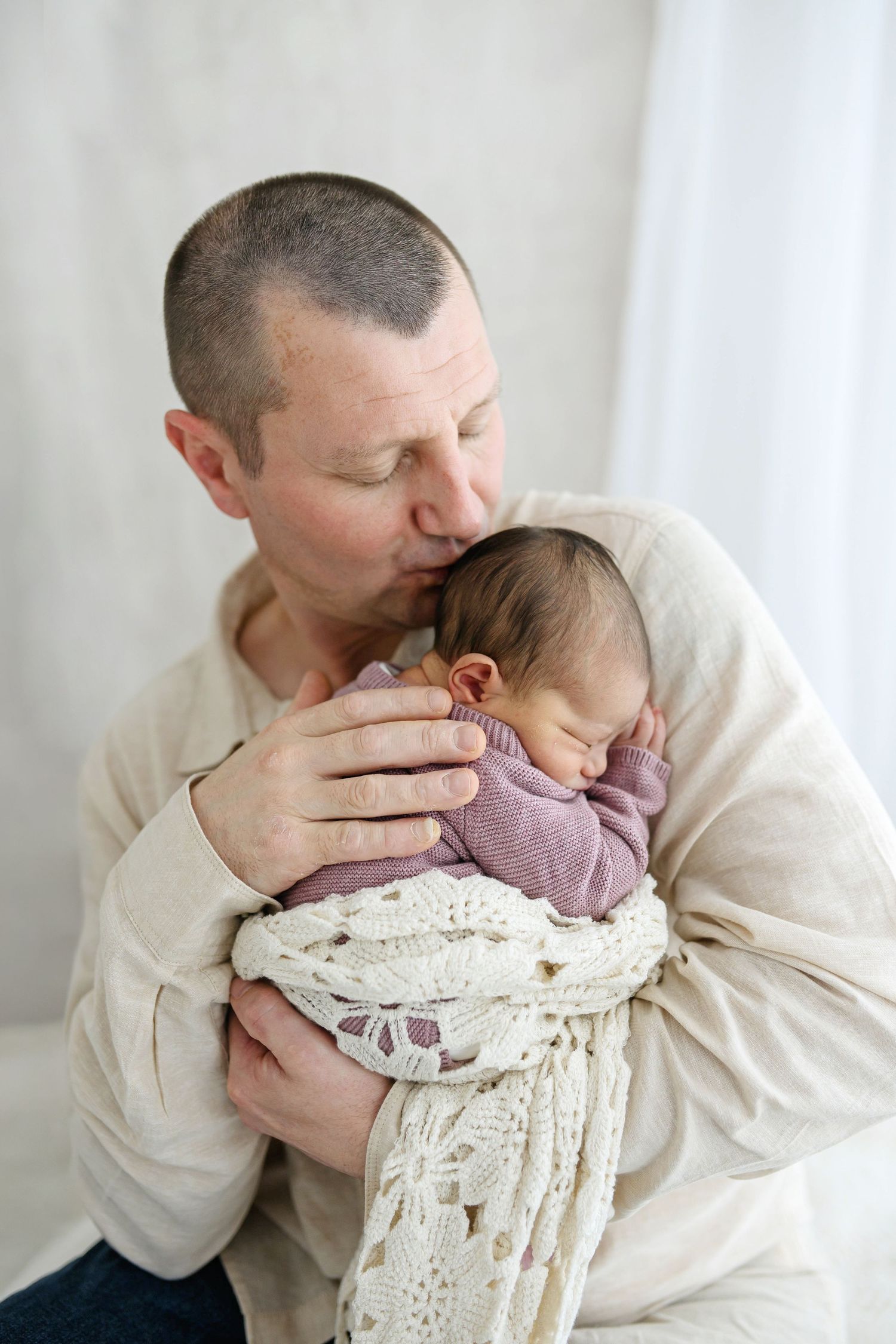 A father tenderly kisses his sleeping newborn baby wrapped in a white lace blanket.