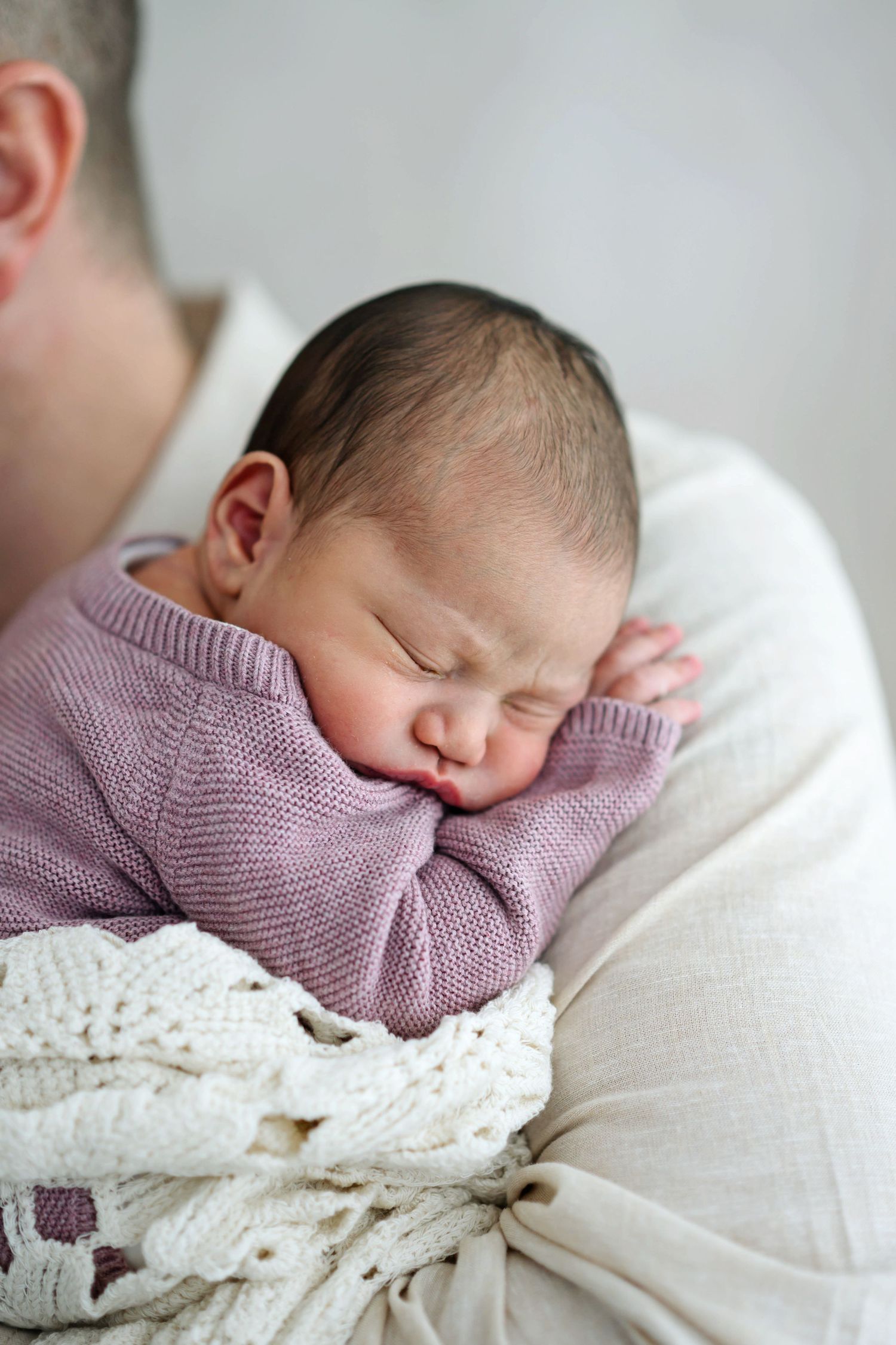 Newborn baby sleeping peacefully in a pink knit sweater, cradled in a parent's arms with a white blanket.