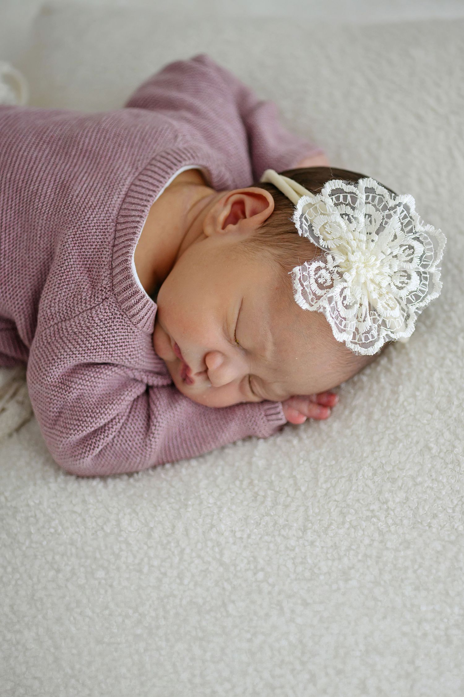 Sleeping newborn baby girl in pink knit outfit with white lace headband bow, lying on soft white blanket.