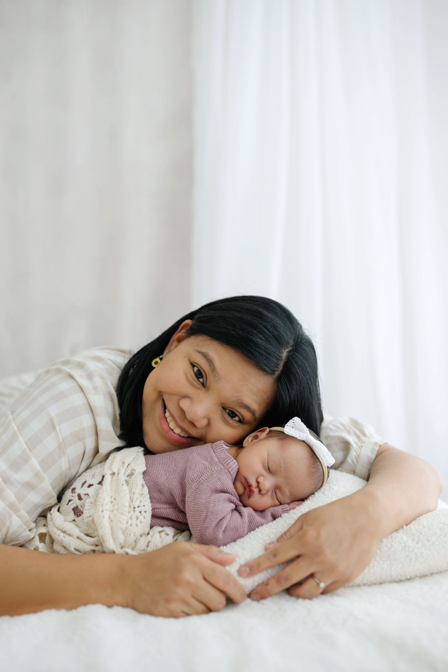 Smiling mother lying on white bed cheek-to-cheek with sleeping newborn baby wrapped in pink outfit.