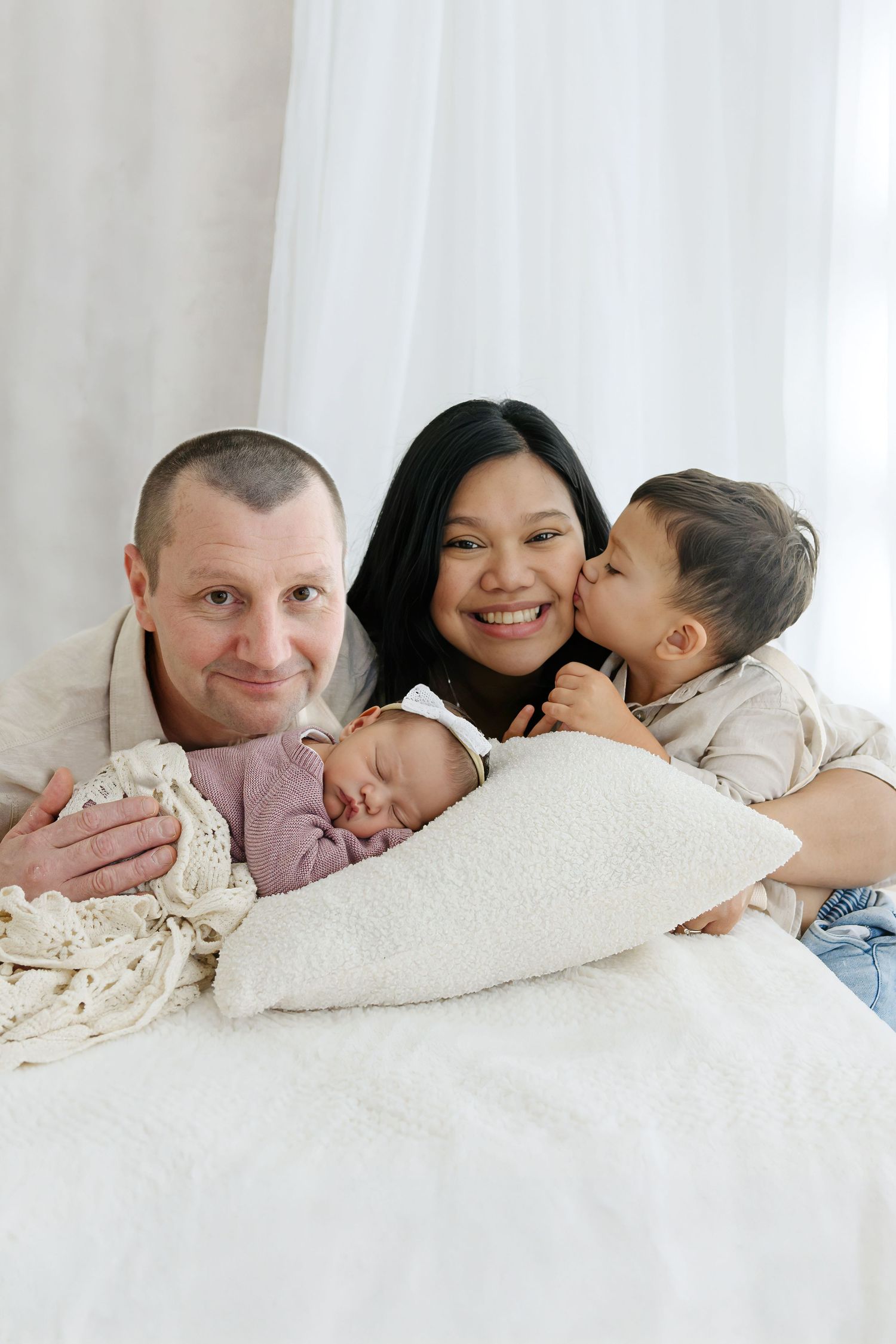 Happy family portrait with parents and two children, including a sleeping newborn baby wrapped in white, on a soft bed.