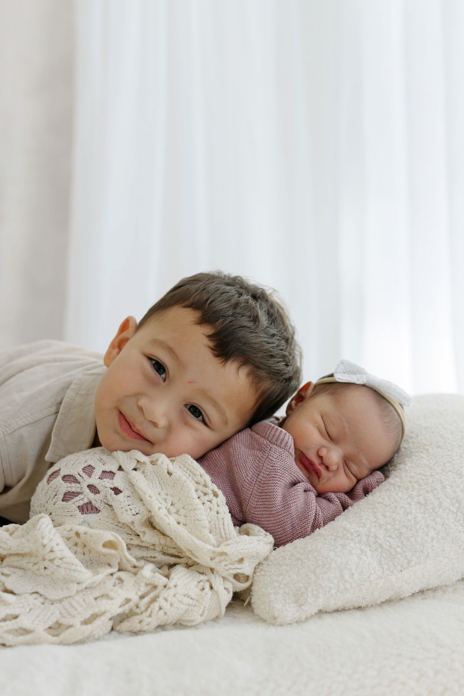 Young boy smiles while resting his head next to sleeping newborn baby sibling wrapped in soft blankets.