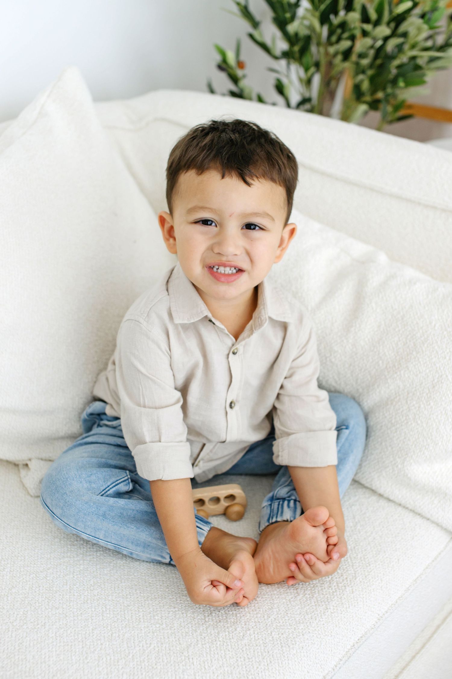 Smiling toddler boy sitting cross-legged on a white sofa, wearing a beige shirt and jeans, holding a wooden toy.