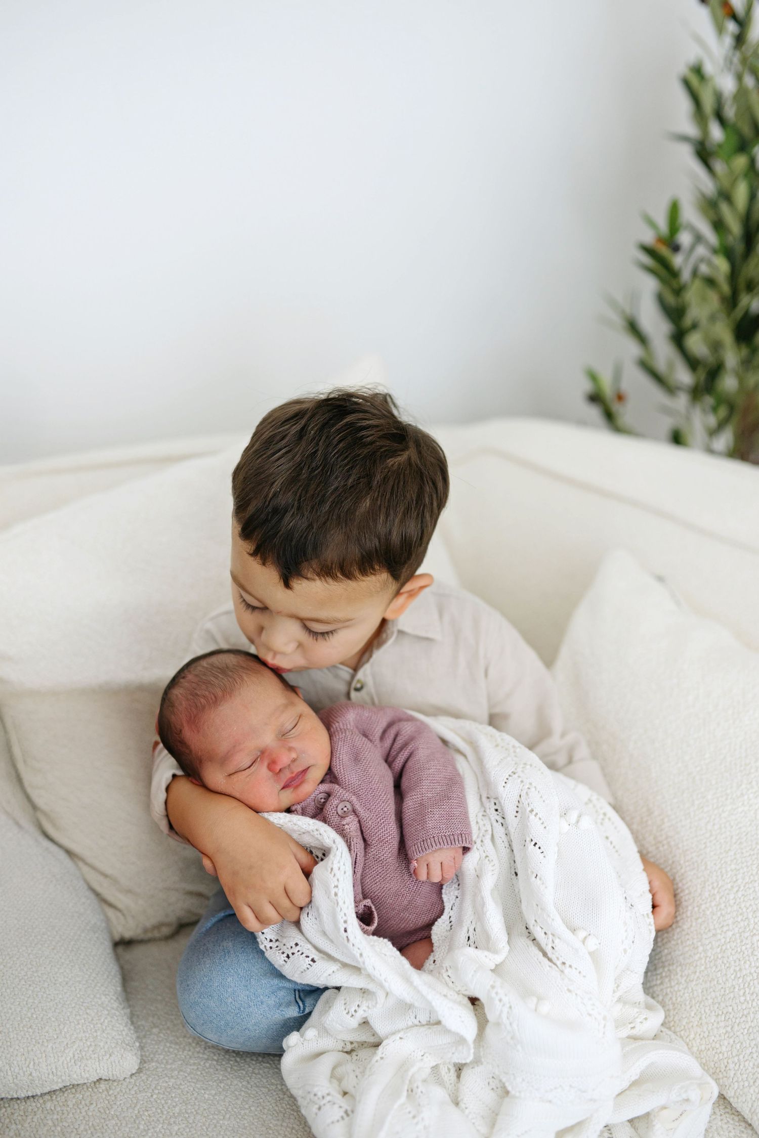 Young boy gently holding sleeping newborn baby sibling wrapped in white knit blanket on cream sofa.