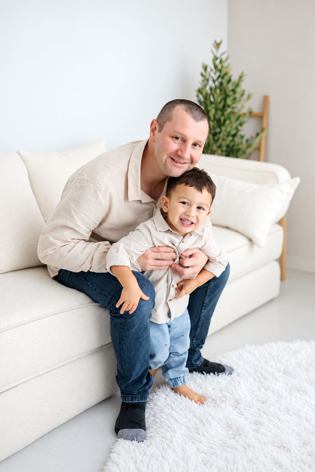 A smiling father sits on a white sofa hugging his young son on a fluffy rug in a bright living room.