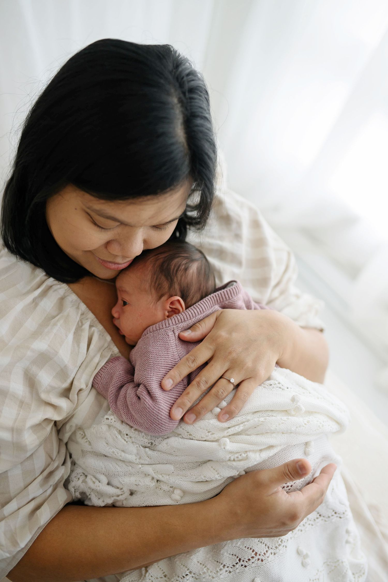 A mother tenderly holds and kisses her sleeping newborn baby wrapped in a soft blanket.