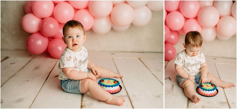 Baby's first birthday cake smash session with colorful rainbow cake and pink balloon backdrop on white wood floor.