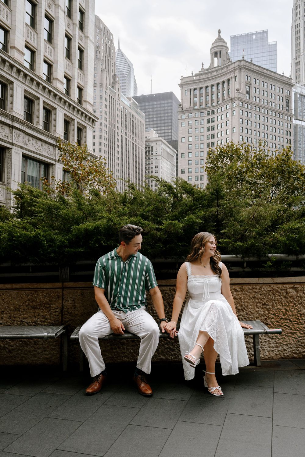 Couple holds hands on a bench in downtown Chicago, wearing white dress and green striped shirt, with city skyline behind.