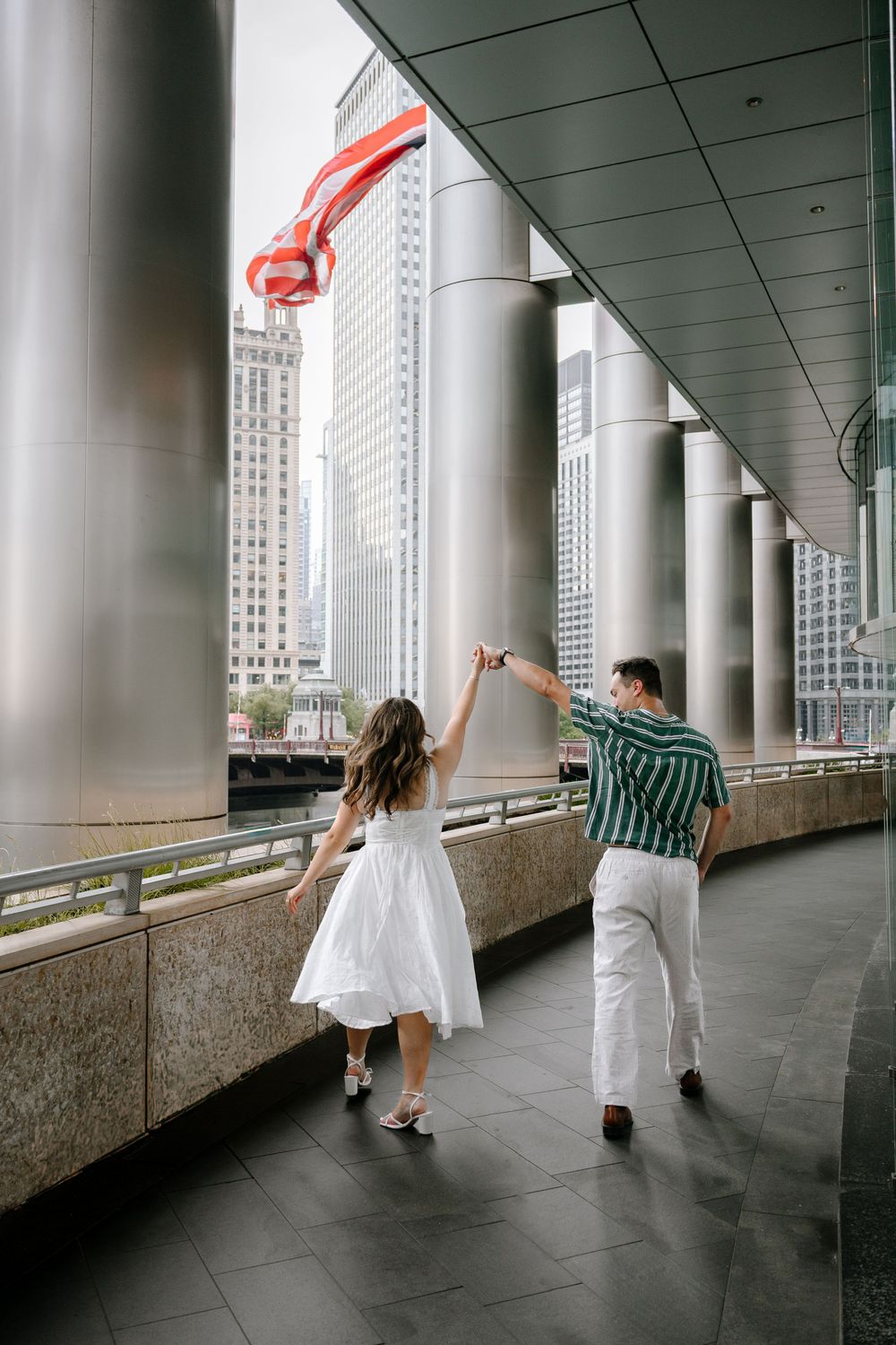 Couple dancing under city colonnade, woman in white dress, man in striped shirt, waving American flag, urban skyline backdrop.
