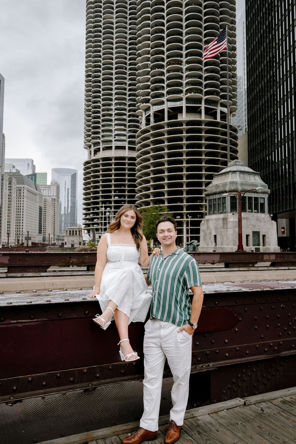 Couple posing on a bridge in Chicago with iconic Marina City towers and American flag in the background.
