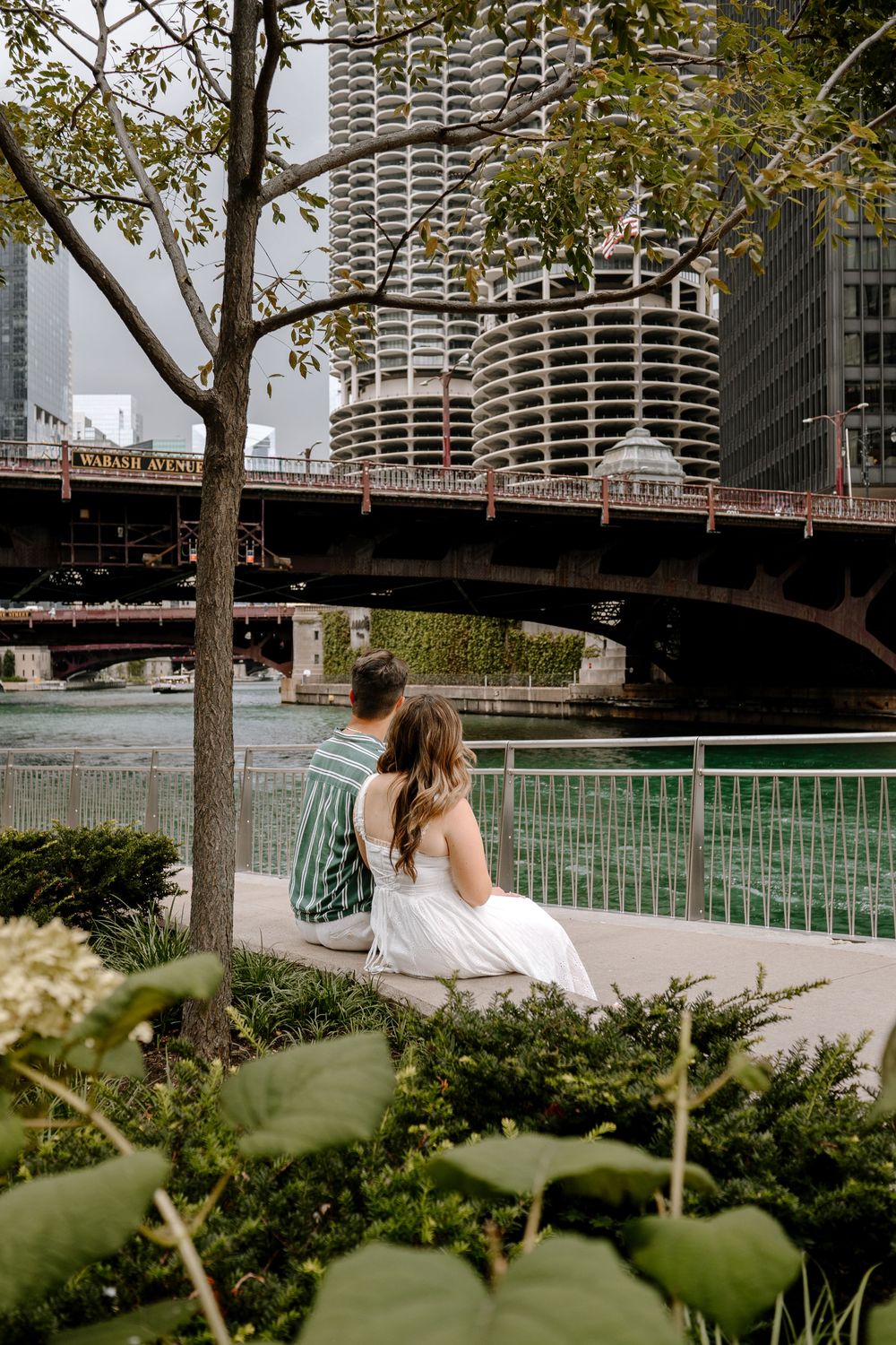 Couple sitting together along the Chicago Riverwalk with Marina City towers and a bridge visible behind them.